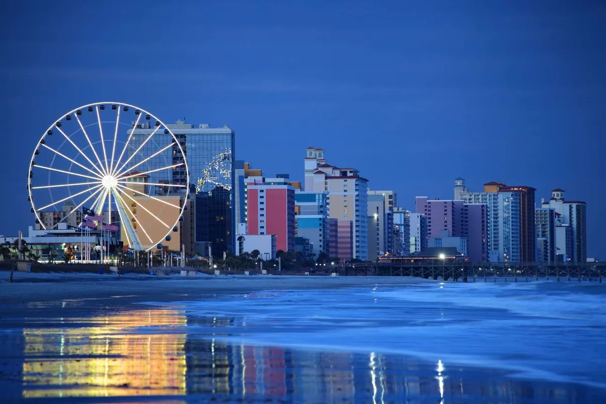 A vibrant scene featuring a Ferris wheel set against the ocean, with colorful beachfront buildings and reflections shimmering on the water. The festive lighting and beachside setting evoke a sense of fun and adventure, inviting guests to explore the lively local attractions just beyond the property.