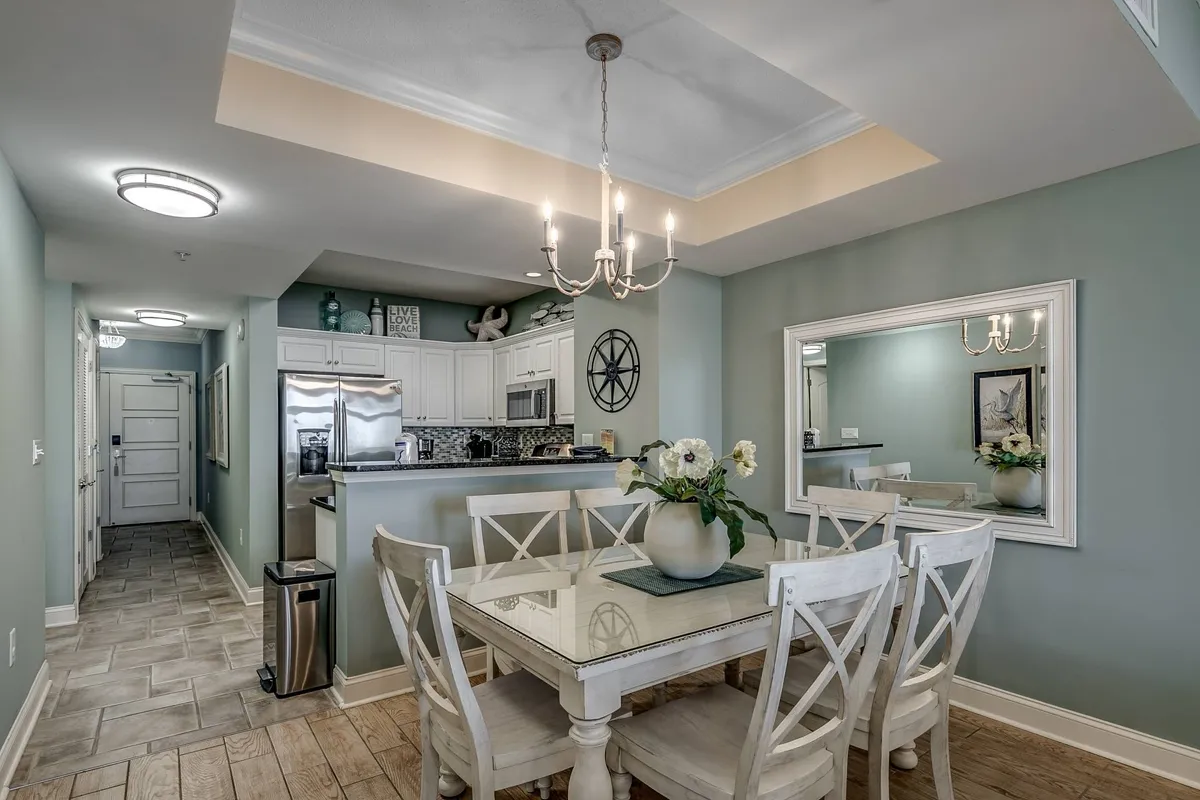 A warm, welcoming dining area centered around a large table, illuminated by a statement chandelier and complemented by a decorative wall mirror. The space is set for gatherings, evoking memories of family dinners and lively group activities.