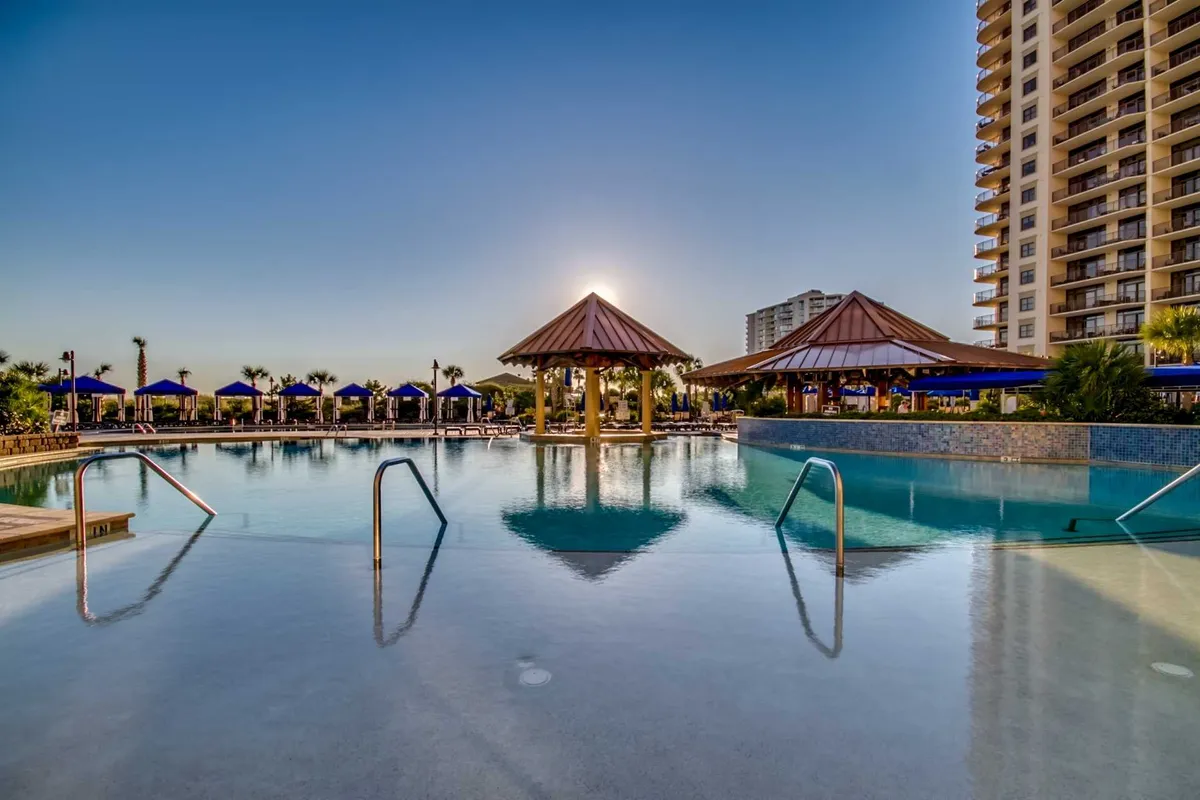 A tranquil sunset scene showcases a swimming pool framed by pavilions, palm trees, and plush lounge chairs. The sun reflects off the water’s surface, creating a picturesque and serene ambiance. The image conveys a sense of relaxation and premium resort living.