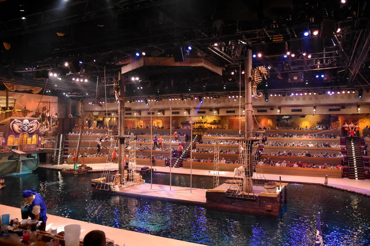 This image captures the vibrant interior of a themed dinner show venue, with a stage featuring water elements, rows of audience seating, and immersive decorations. The atmosphere is lively and energetic, hinting at interactive performances and dining experiences.