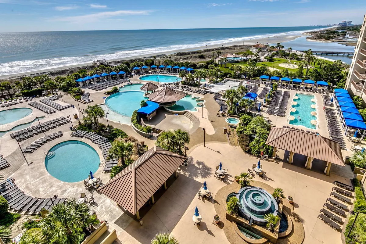 This image presents an expansive view of a pool complex set against a beachfront backdrop. Multiple swimming pools are surrounded by palm trees, lounge chairs, and inviting cabana structures, evoking a lively, vacation-like atmosphere. The scene suggests ample space for both relaxing and socializing, with the beach and ocean visible just beyond the pools.
