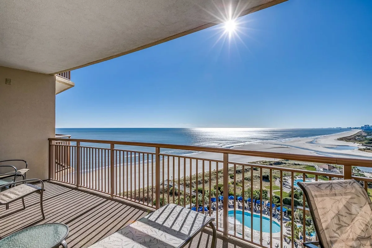 A stunning ocean view from a private balcony, framed by a sleek railing and bathed in sunlight. The scene features a tranquil expanse of blue water, with inviting spaces to sit and enjoy coffee or conversation while soaking up panoramic views.