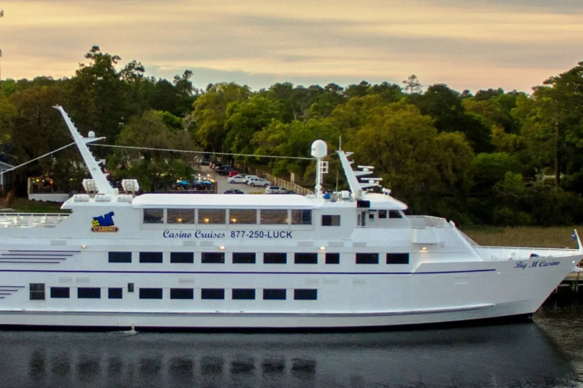 The scene displays a large white yacht branded with 'Casino Cruises,' docked against a lush, green backdrop. The yacht is set up for onboard dining and entertainment, suggesting opportunities for group gatherings, sunset watching, and scenic cruises.