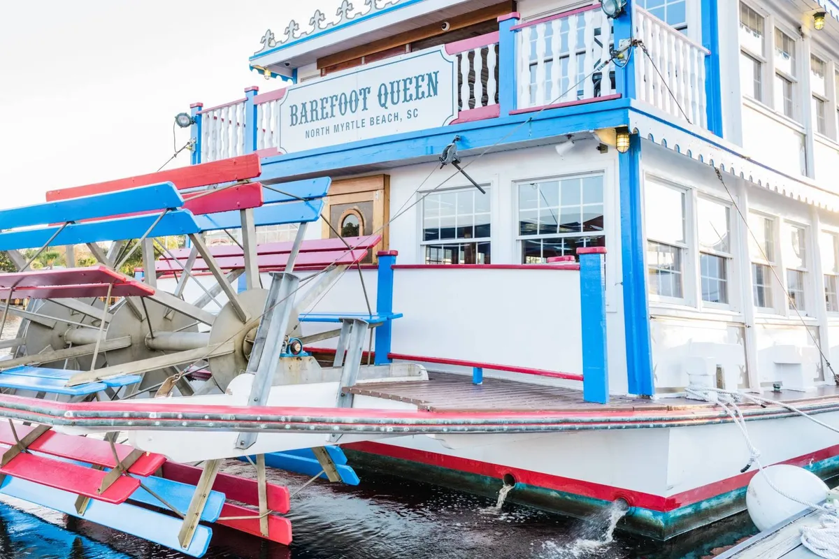 This image features a brightly painted restaurant with a colorful paddle wheel overlooking the water. The restaurant's name is visible on a sign, and the lively colors and architecture evoke a cheerful, welcoming atmosphere. Outdoor dining and the proximity to the water hint at social gatherings and the vibrant experience available at Barefoot Landing.